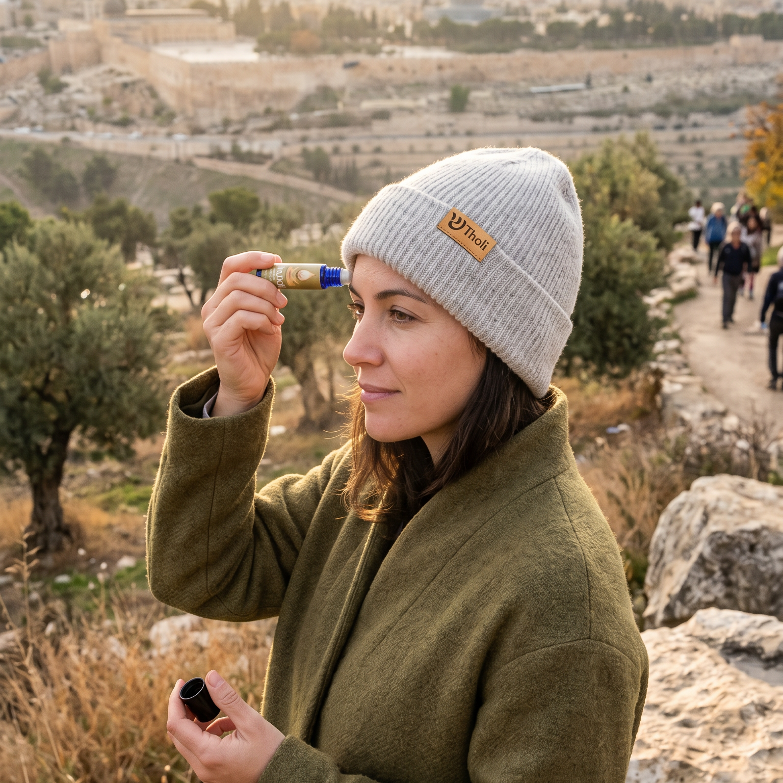 A woman wearing an olive green coat and a light gray beanie stands outdoors in Jerusalem, holding a small blue and yellow item near her face. The background shows a scenic view of the city with the Dome of the Rock under a warm sunset sky.